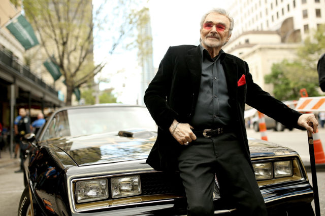 Burt Reynolds leaning against the front of a car