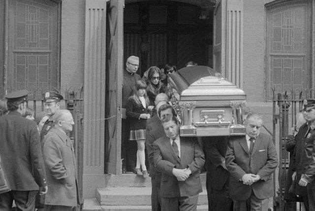 Casket carried by a group of men in suits walk out of the doors of a church followed by the family.