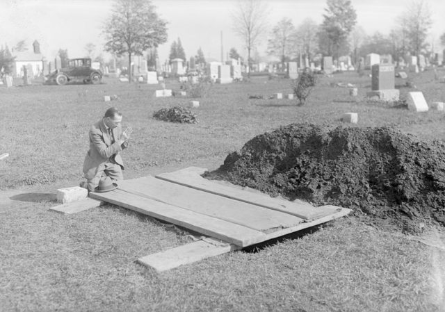 Jimmy Mills kneeling in prayer beside a freshly dug grave covered with wood.
