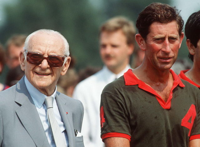 Armand Hammer in a grey suit and large sunglasses stands beside Prince Charles in a green and red polo shirt.