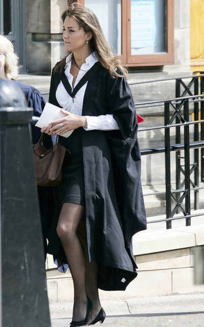 Kate Middleton walking while wearing a black graduation gown over a black skirt and white shirt.
