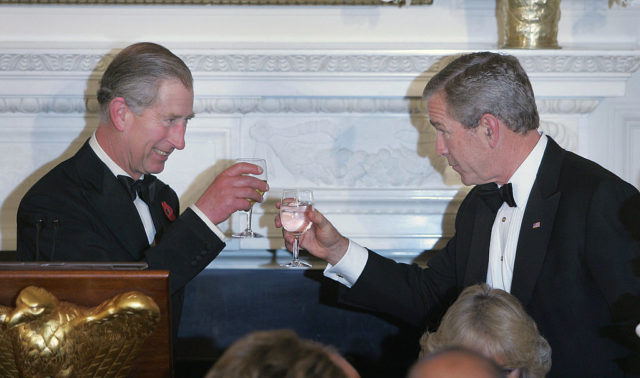 King Charles III and President Bush wearing suits clink wine glasses.