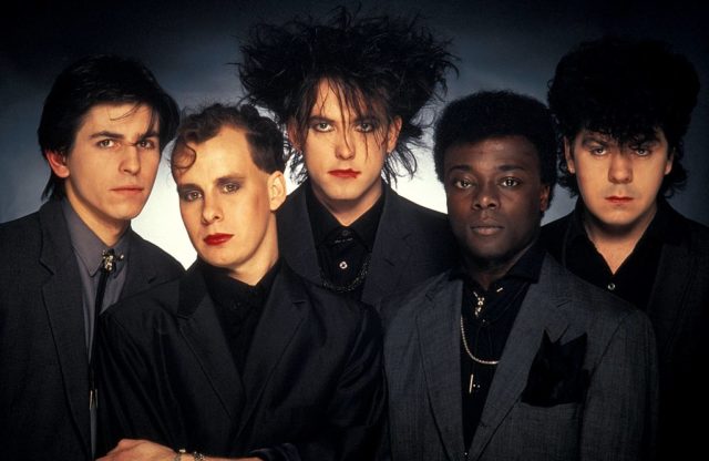 Members of the band 'The Cure' wearing black suits with Robert Smith standing in the middle.