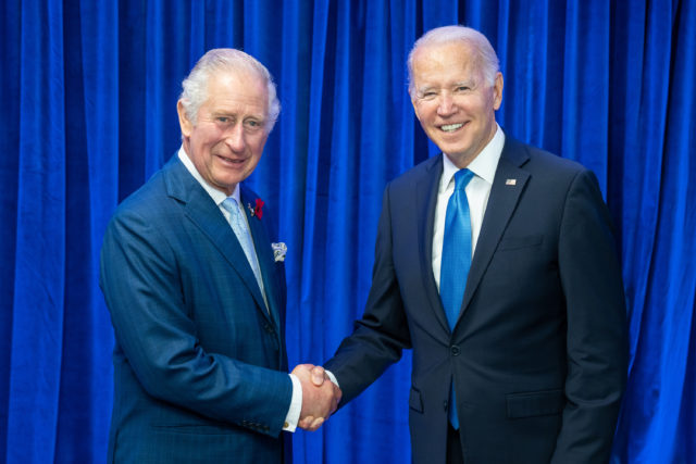 King Charles III and President Biden shaking hands in front of a blue curtain.