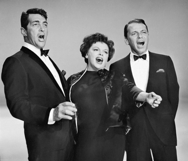 Dean Martin and Frank Sinatra in black suits singing with Judy Garland in a black dress.