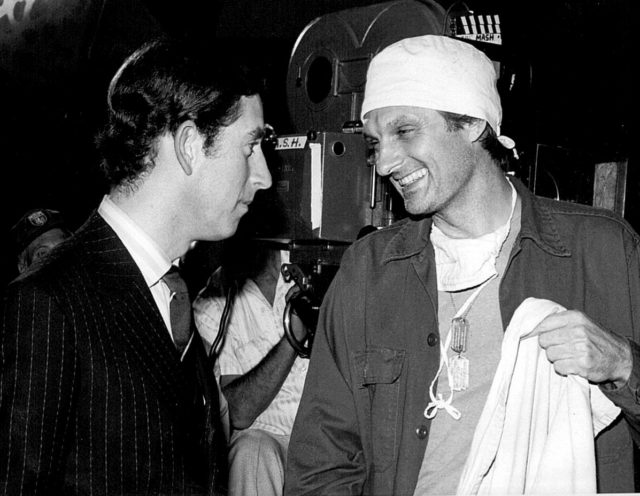 Alan Alda wearing scrubs smiles while he talks to Prince Charles.