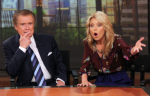 Regis Philbin and Kelly Ripa sitting at a wooden table while Kelly waves her hand around.