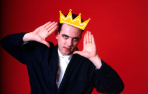 Robert Smith with his hands framing his face in front of a red back ground, with a cartoon crown on his head.