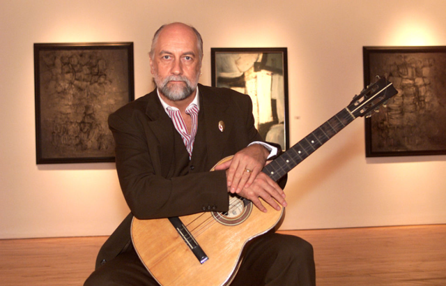 Mick Fleetwood wearing a suit scowling with a guitar in his lap, paintings in the background.
