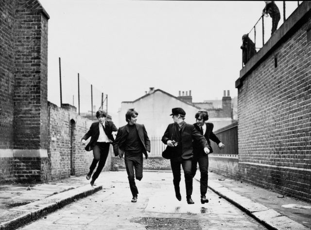 The Beatles running down a street while looking behind them wearing suit jackets. 