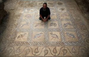 Farmer crouching on a detailed mosaic with birds, looking up at the camera.