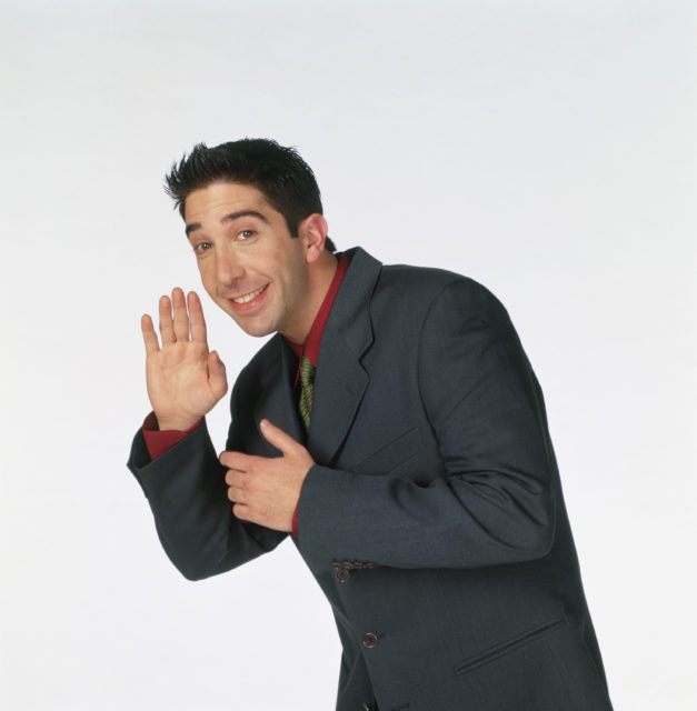 David Schwimmer as Ross Geller wearing a grey suit and waving at the camera.