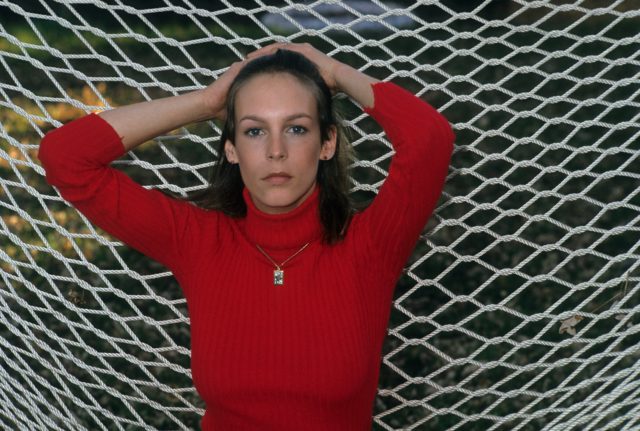 Headshot of a young Jamie Lee Curtis with his hands on her head in a red turtleneck