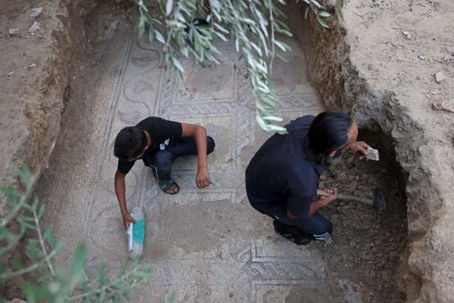 Man and his son, both wearing black, crouch on a mosaic in the middle of a dirt hole and dig out the sides.