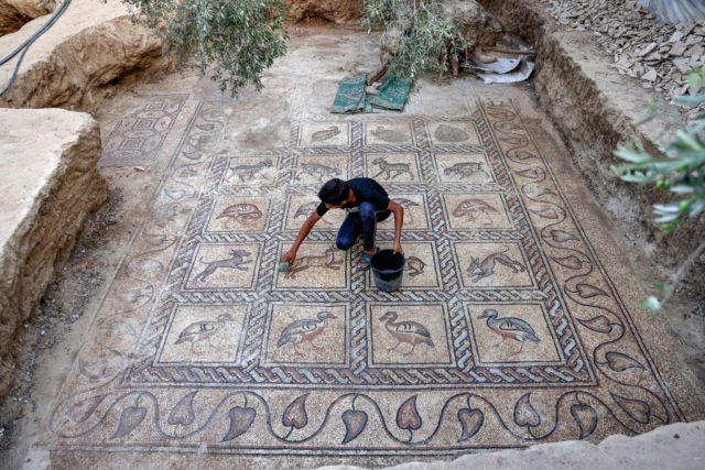 Young man with a bucket and sponge crouching to clean a mosaic with a variety of birds and animals on it.