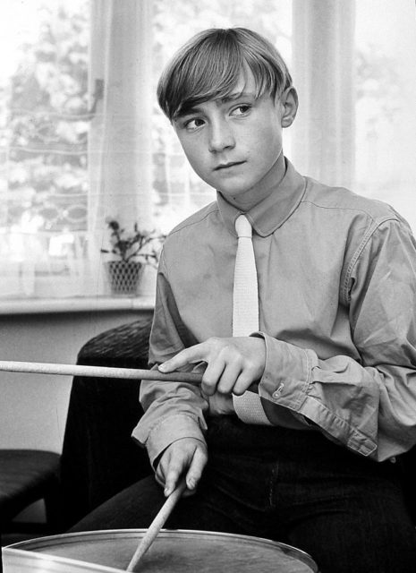 Young Phil Collins wearing a collared shirt and tie, holding drum sticks. 