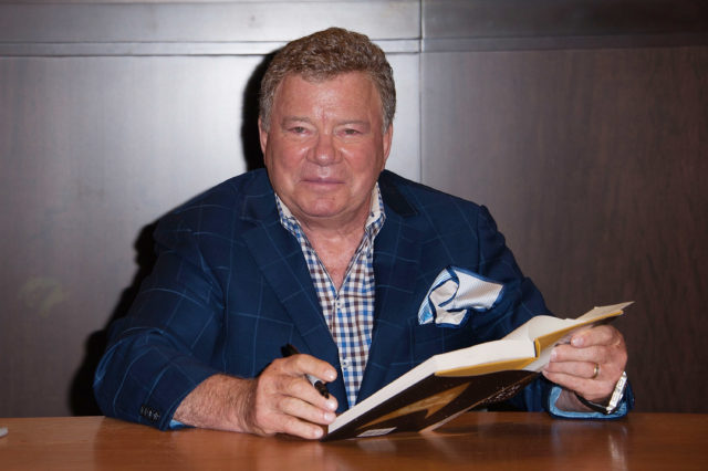William Shatner sitting at a desk holding a book