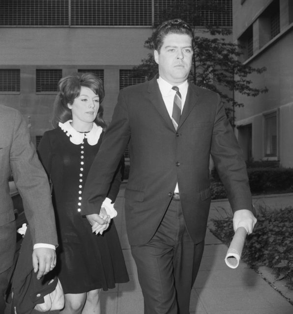 A man in a suit walks in front of a woman wearing a black dress with a white collar and buttons