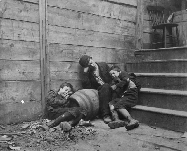 Three children sleep huddled against a wooden barrel in the street