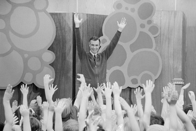 Mr. Rogers on a stage in front of a group of children with their hands in the air, also with his hands in the air.
