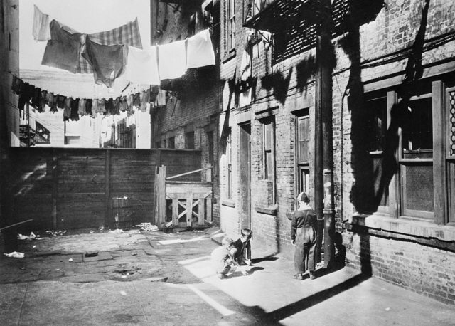 Young children play marbles in the back of a building