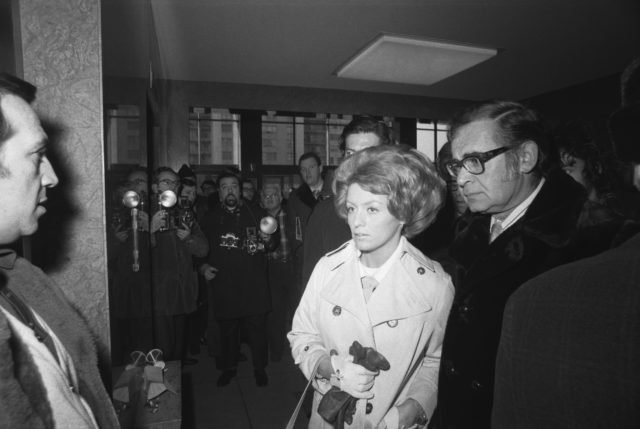 A woman walks with a man in a room filled with photographers and reporters in the foreground
