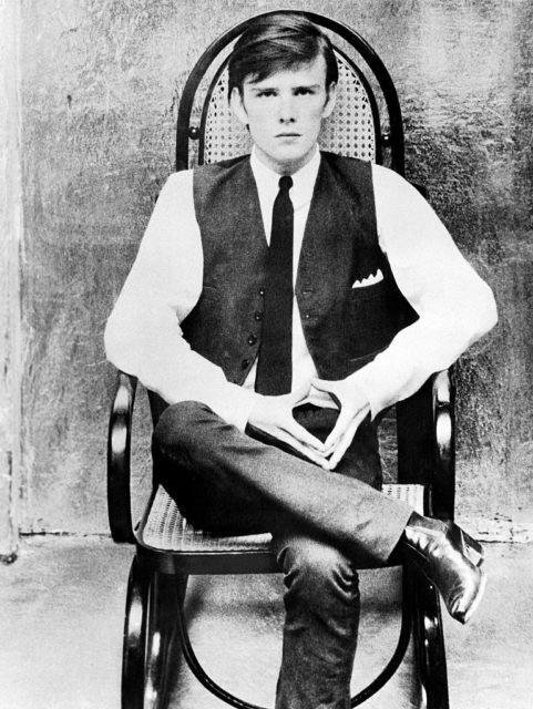 Stuart Sutcliffe wearing a waist coat, while shirt and tie, sitting in a chair with his leg crossed over his knee and his hands clasped together.