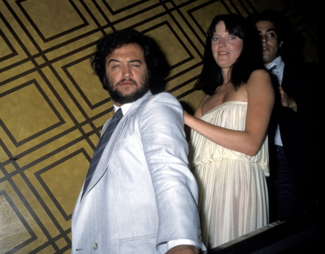 John Belushi, his wife Judy behind him, walking down a staircase in a blue suit jacket