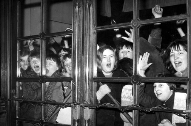 Young actors and actresses playing Beatles fans, screaming against a glass window.