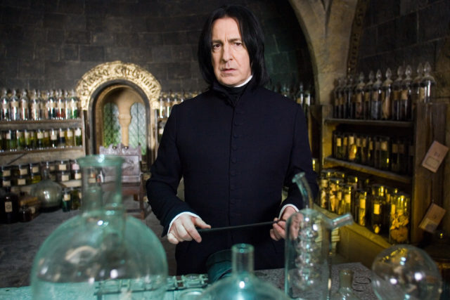 Alan Rickman in a black robe with black hair as Severus Snape holding a wand in front of a row of glass bottles.
