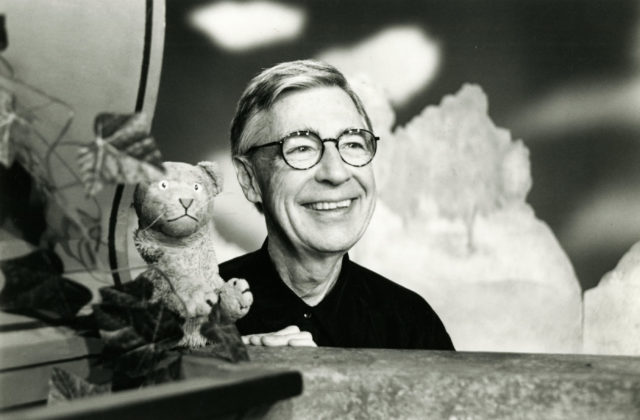 Mr. Rogers wearing glasses smiling while sitting beside a puppet.