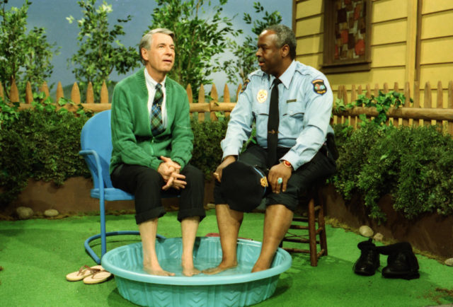 François Scarborough Clemmons dressed as a security guard, and Mr. Rogers in a green sweater, sit beside each other with their feet in a kiddy pool.
