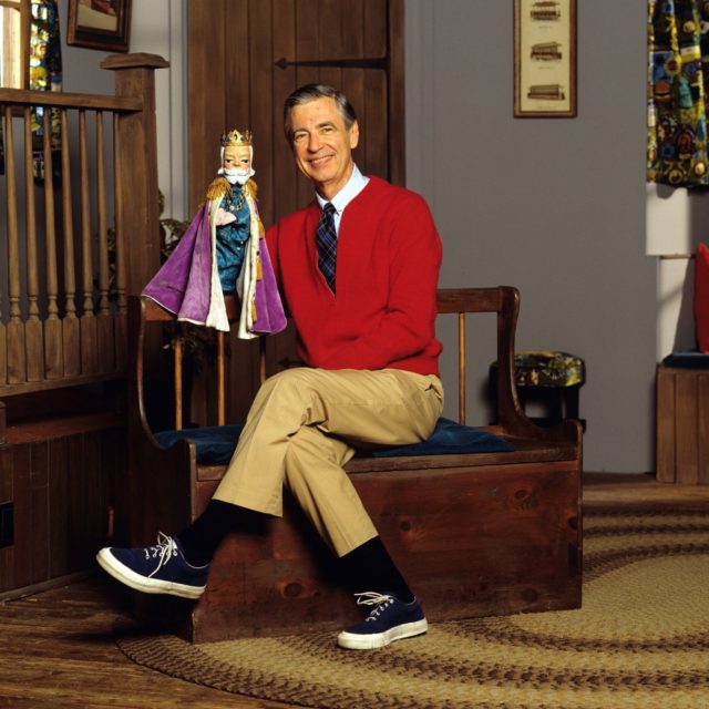 Mr. Rogers sitting in tan pants and a red sweater with a puppet on one hand. 