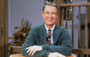 Mr. Rogers in a green cardigan and a tie, with his arms crossed smiling at the camera.