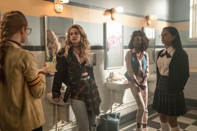 Lili Reinhart as Betty Cooper leaning against a sink in the girls bathroom surrounded by other girls, wearing a leather jacket and fingerless gloves.