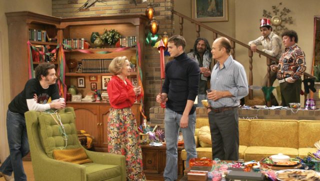Debra Jo Rupp as Kitty Forman in the living room surrounded by many of the cast while wearing a read blouse and floral skirt.