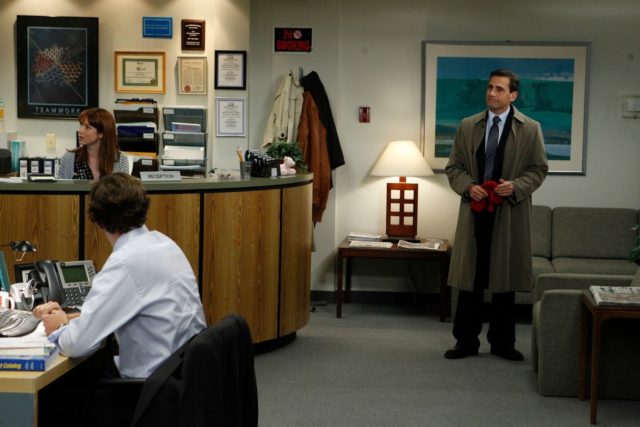 Steve Carell as Michael Scott standing by the reception desk in the office holding a pair of red mittens.
