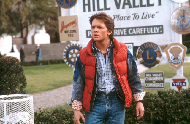 Young Michael J. Fox as Marty McFly wearing jeans and a jean jacket with a red vest overtop.