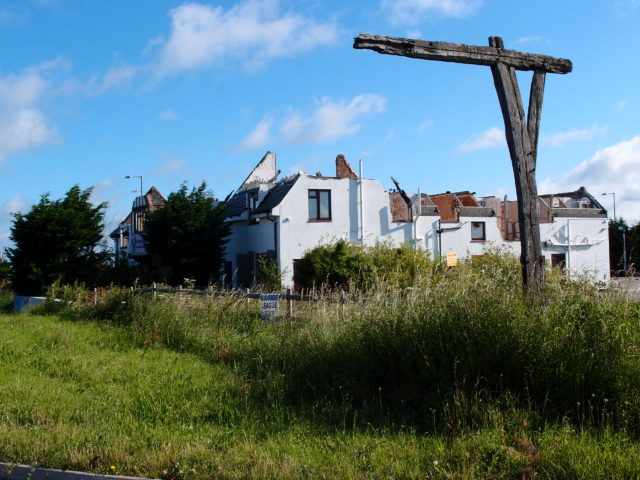 A wood gibbet stands in a field, a house in the background