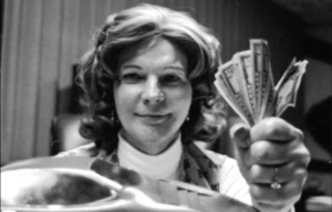 Elizabeth Carmichael holding a fistfull of money with a large diamond ring on her hand while staring into the camera.