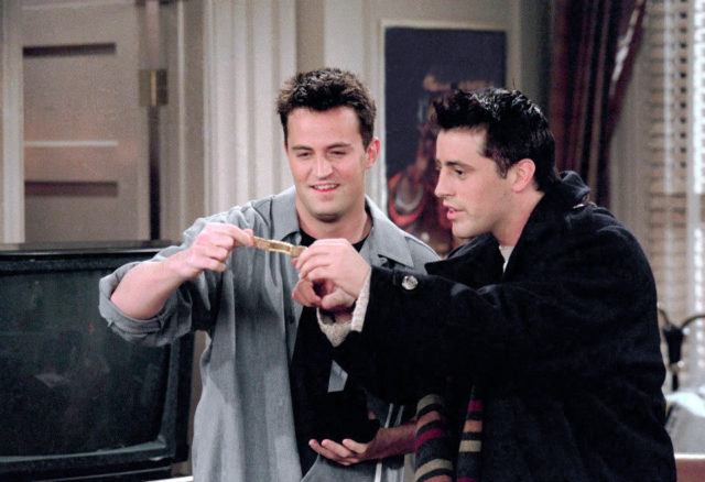 Matthew Perry as Chandler Bing in a grey collared shirt, and Matt LeBlanc as Joey Tribbiani in a black jacket, holding a note between them.