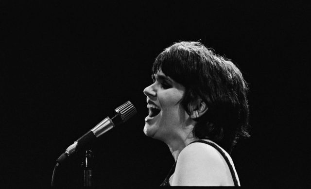 Young Linda Ronstadt with short dark hair in profile while singing into a microphone.