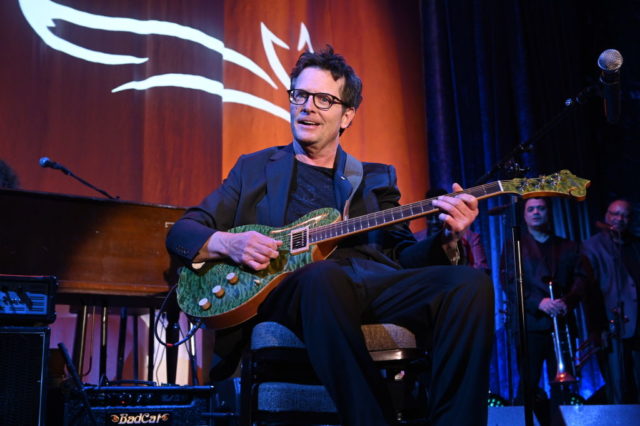 Michael J. Fox sitting on a chair playing a green guitar.