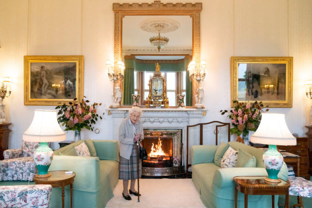 Queen Elizabeth wearing a plaid skirt and grey cardigan stands in front of a lit fire in the middle of a sitting room.