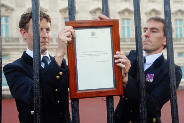Two men wearing black jackets, ties, and white collared shirts reach through the gates at Buckingham Palace to post a death notice of the Queen.