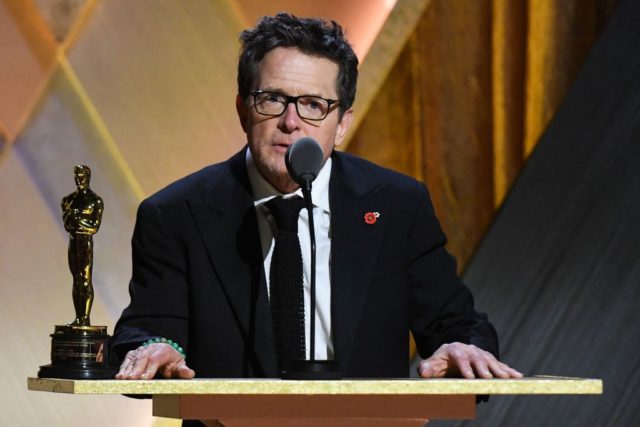 Michael J. Fox in a black suit with white shirt talking into a microphone with an Academy Award on a podium beside him. 