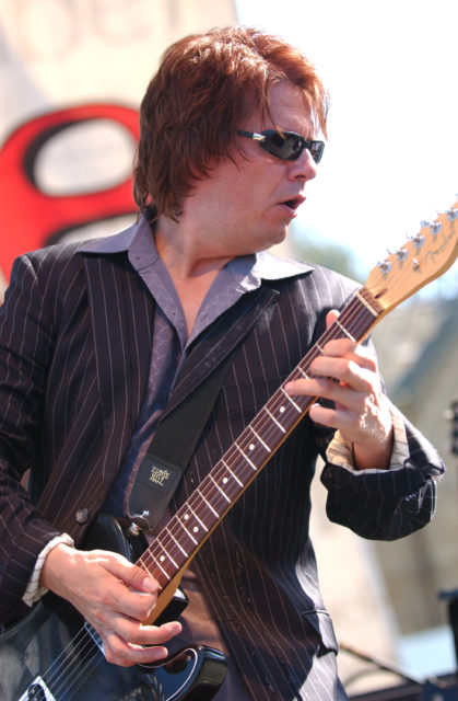 Andy Taylor is a suit jacket and sunglasses playing the guitar