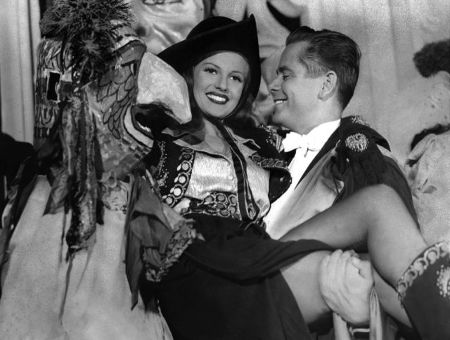 Glenn Ford carrying Rita Hayworth in his arms while she wears an elaborate costume with a pirate hat.