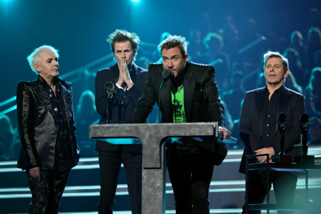 Four members of Duran Duran speaking up at a podium on a stage