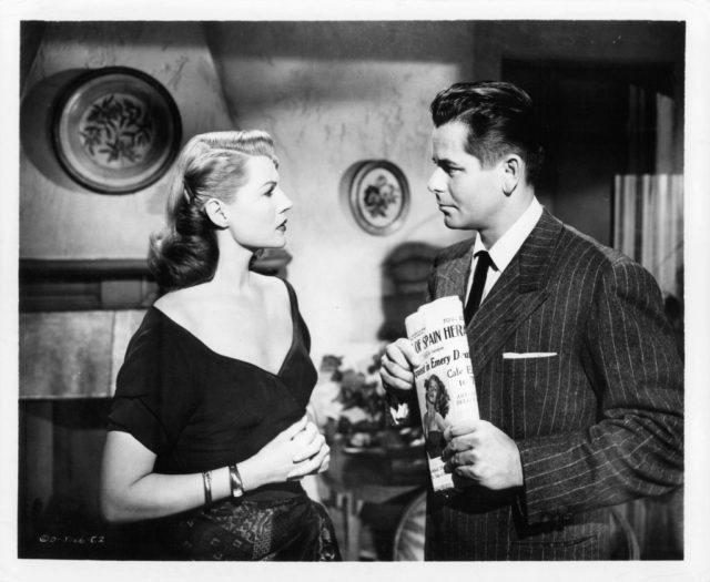 Rita Hayworth, in a black dress, and Glenn Ford in a dark pinstripe suit holding a newspaper, stare at each other intently.